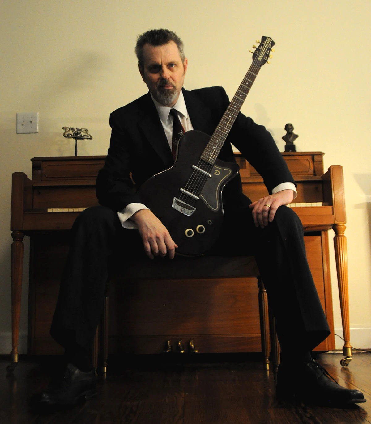 Dramatic photo of Chris Black wearing a dark suit and a blood red tie with an electric guitar sitting with his back to a piano. 